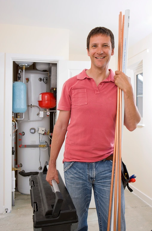 Smiling plumber in a red polo shirt holds copper pipes and a toolbox, standing in a bright room by a water heater. The atmosphere is friendly and professional.