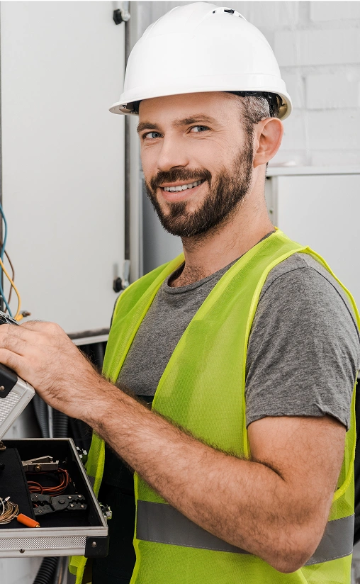 Smiling electrician wearing a white hard hat and neon safety vest works on a control panel, holding tools.