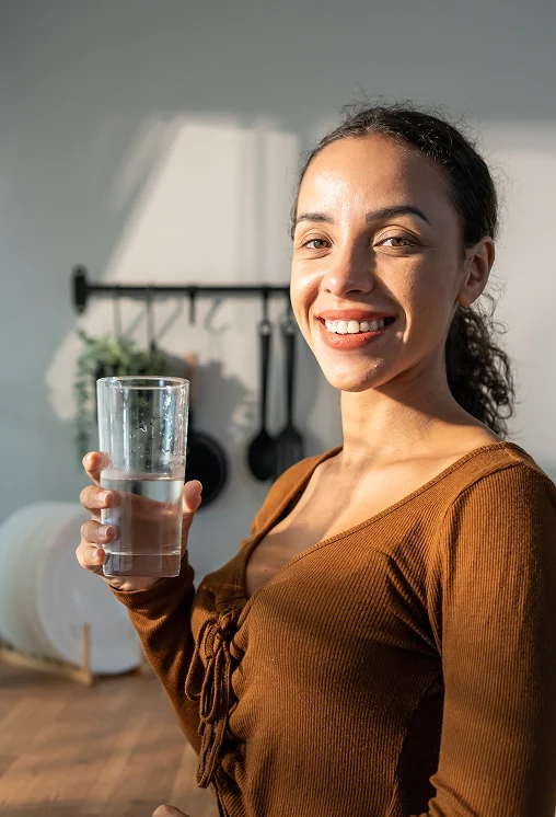 A woman in a brown top smiles while holding a glass of water in a sunlit room, creating a warm and cheerful atmosphere. Kitchen utensils hang in the background.