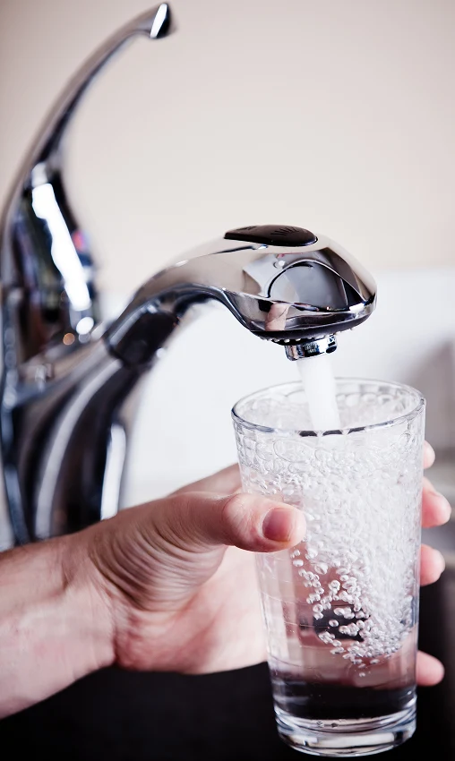A hand holds a clear glass under a modern silver faucet, filling it with bubbly, running water. The setting is a clean, well-lit kitchen.