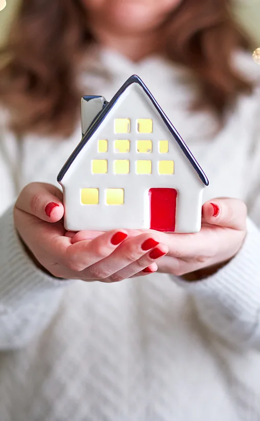 A woman in a white sweater holds a small, ceramic house with glowing yellow windows and a red door. Her nails are painted red, creating a cozy vibe.