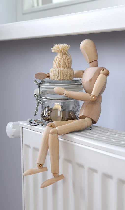 Wooden mannequin sitting on a radiator, holding a jar with a knitted hat. The scene is cozy and whimsical, with neutral tones and a warm feel.