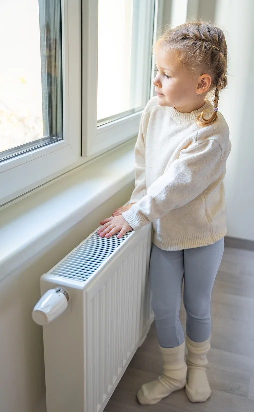 Child standing near a home radiator and feeling warm indoor heating