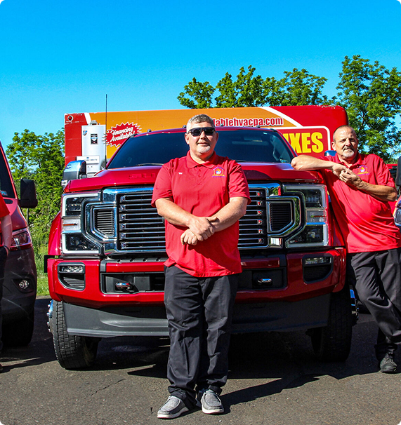 Two men in red shirts stand cheerfully beside a large red truck with a vibrant advertising banner, set against a backdrop of green trees.