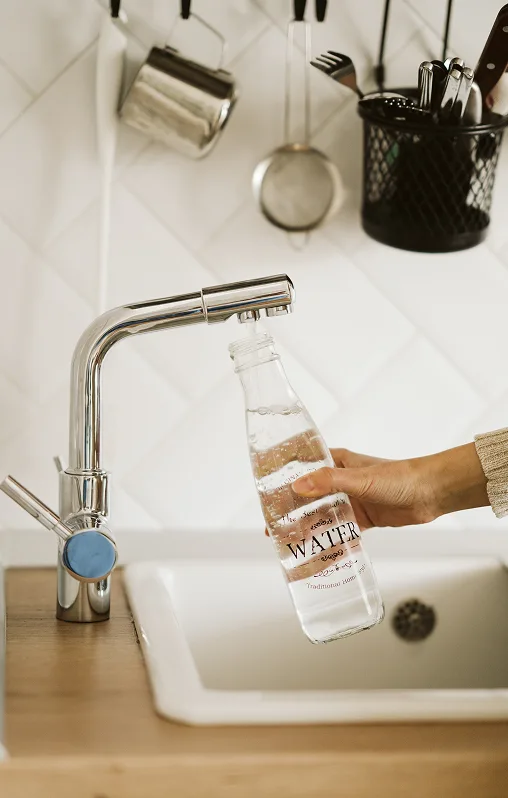 A hand fills a glass bottle labeled "Water" from a modern kitchen faucet. The background shows hanging utensils and a tidy sink area, conveying a clean and organized atmosphere.