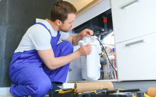 A plumber in blue overalls kneels, focused, fixing pipes under a kitchen sink. Tools and a toolbox are scattered nearby. The scene is industrious and attentive.