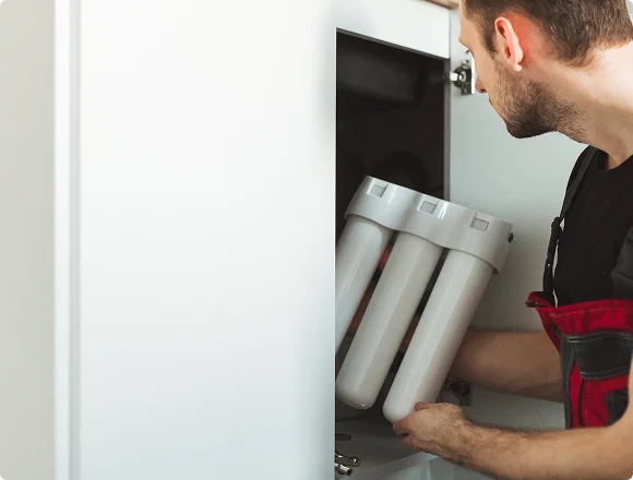 A person in a black shirt and red overalls installs a water filtration system under a white kitchen counter, focusing intently on the task.