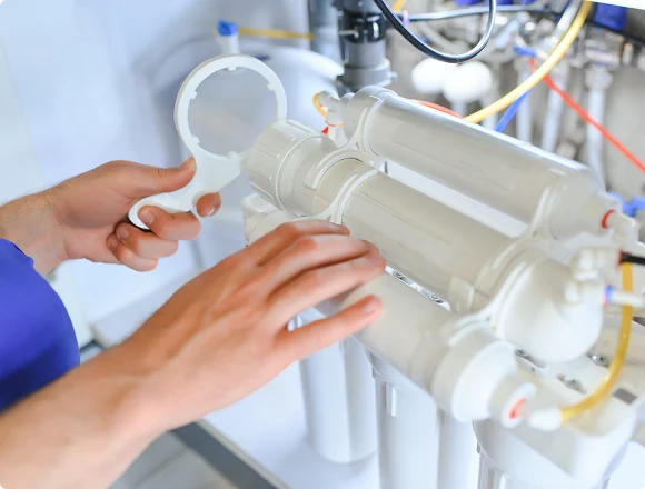 Hands inspect a complex water filtration system with multiple white cylindrical filters and yellow tubing, suggesting maintenance or installation.