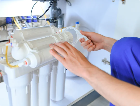 Hands installing a water filtration system, focusing on white cylindrical filters beneath a sink. The scene conveys a sense of precision and concentration.