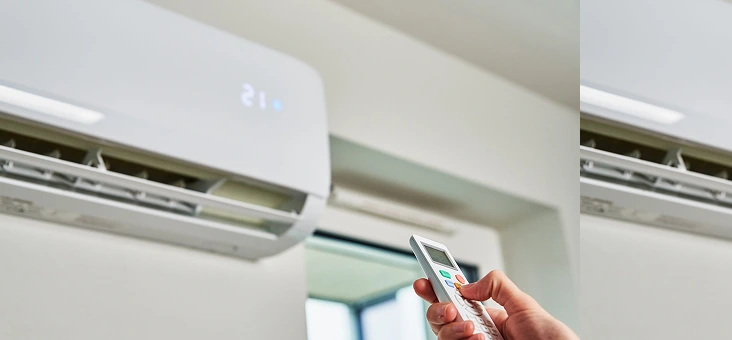 A person holds a remote control, pointing it at a wall-mounted air conditioner set at 21°C. The setting appears in blue, in a bright room.