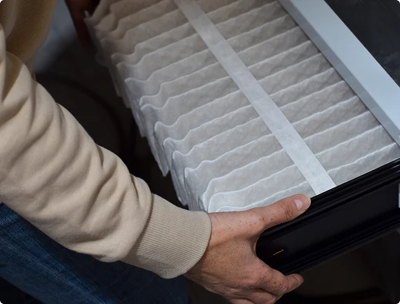A person in a beige sweater replaces a pleated white air filter, carefully fitting it into a black slot. The scene conveys attention to maintenance.