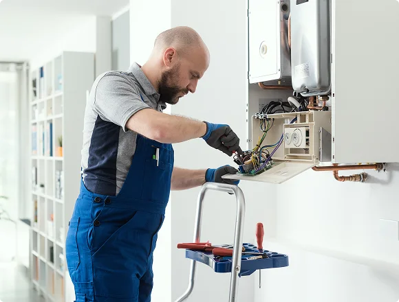 A man in blue overalls and gloves repairs a wall-mounted boiler in a bright room. He uses tools from a nearby step ladder with a focused expression.