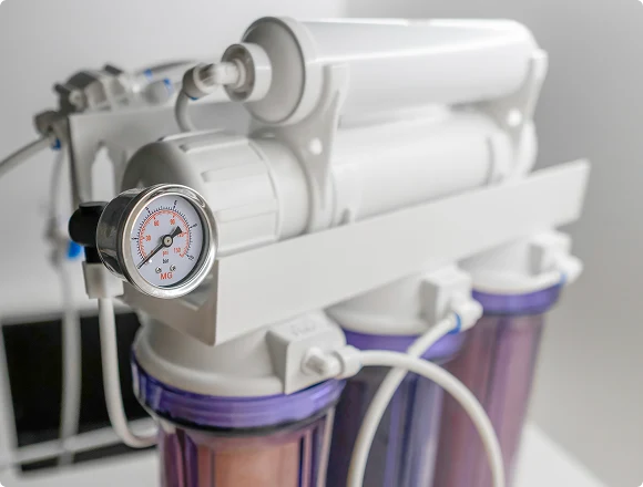 Close-up of a white water filtration system with three purple-tinted canisters and a pressure gauge. The setting is clean and modern.