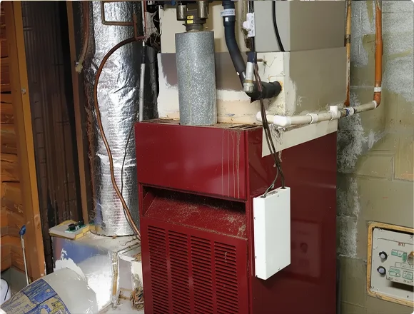 Red furnace in a basement with metal piping and insulation surrounding it. The setting appears functional and industrial, with a utilitarian tone.