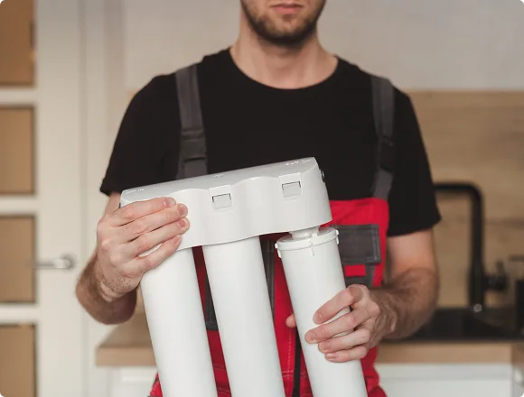 A person in a red and black uniform holds a white water filtration system with multiple cylindrical filters, in a modern kitchen setting.