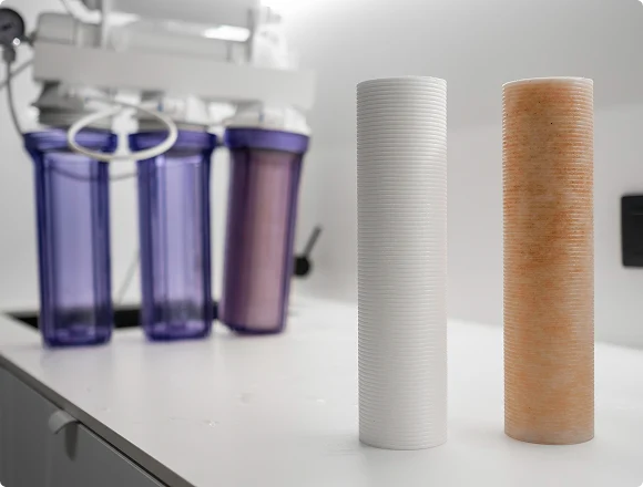 Two cylindrical water filters stand on a white surface. One is clean and white, the other used and brown. Purple filter housings are blurred in the background.