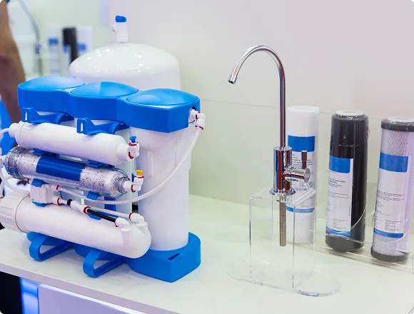 A water filtration system with blue and white components beside a shiny, curved faucet on a countertop. Three filter cartridges are lined up next to it.
