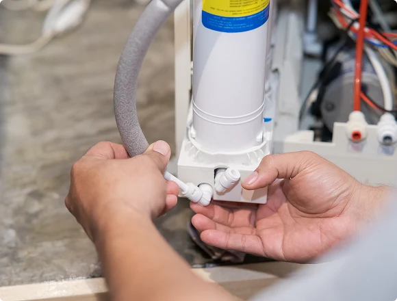 Hands connecting a hose to a water filter unit in a kitchen. The focus is on the precise fitting of the components, highlighting a routine maintenance task.