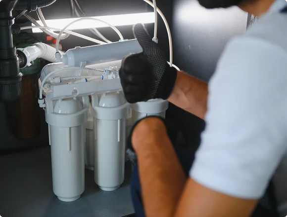 A technician in gloves gives a thumbs-up near an under-sink water filter system with multiple white canisters, highlighting successful installation.