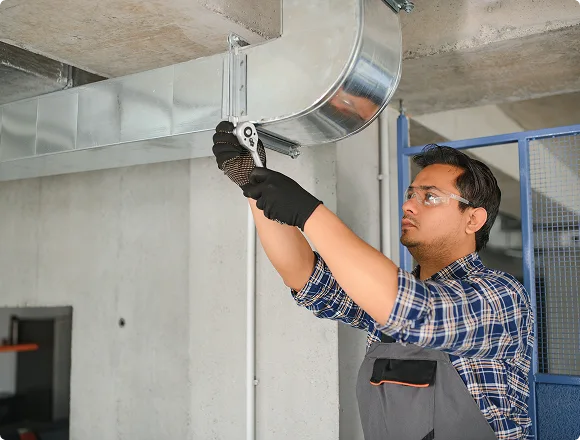 A technician in protective gear tightens a metal duct in an industrial setting. He's focused, wearing gloves and safety goggles, showcasing precision work.