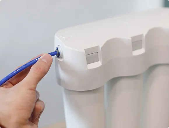 A hand inserts a blue hose into a white water filtration system, suggesting setup or maintenance. The setting is bright, conveying a clean, efficient tone.