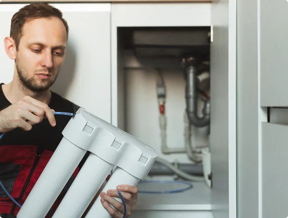 A man installing a water filter under a kitchen sink. He examines the white cylindrical filter, focusing on connecting a blue tube. The scene conveys concentration and care.