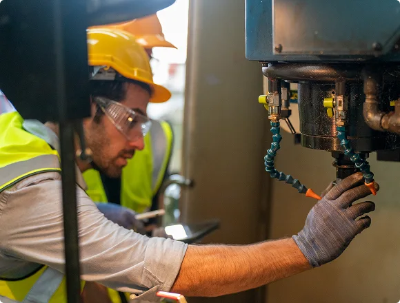 A worker wearing a yellow hard hat, safety goggles, and a reflective vest operates machinery in an industrial setting, displaying focus and precision.