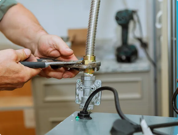 A person is using pliers to fix a metal gas pipe, with a drill in the blurry background. The scene conveys focus and mechanical repair work.
