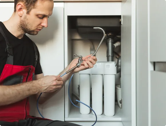A plumber in a red and black uniform fixes a water filter system under a sink. The scene conveys concentration and technical expertise.