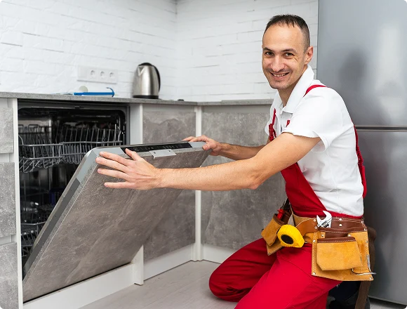Smiling technician in red overalls kneels beside an open dishwasher in a modern kitchen, displaying a screwdriver in a tool belt, conveying readiness.