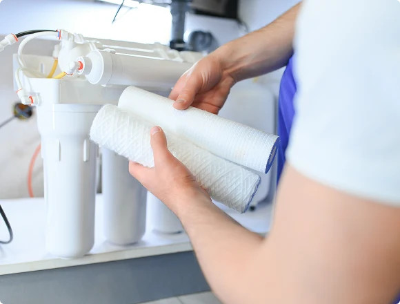 Person replacing water filter cartridges in a filtration system. Hands hold cylindrical filters, suggesting maintenance and clean water assurance.
