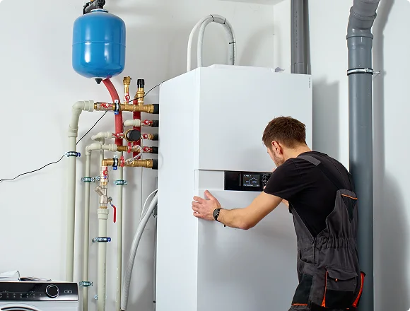 A technician in a black uniform checks a large white boiler system. Surrounding pipes and a blue tank are visible, indicating a maintenance task.