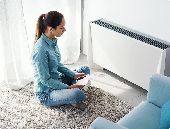 A woman sits cross-legged on a beige carpet, using a tablet. She's near a modern white heater, in a bright room with sheer curtains, creating a cozy atmosphere.