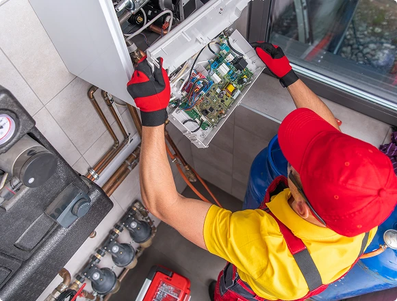 A technician in red and yellow uniform and gloves works on an open boiler panel, surrounded by tools and pipes, conveying focus and precision.