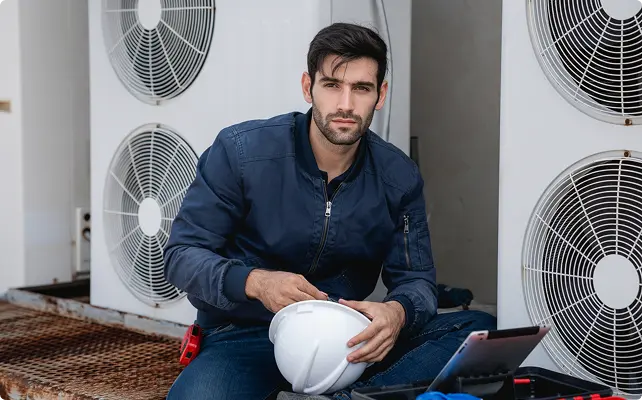 Technician sitting near outdoor HVAC units holding a hard hat.