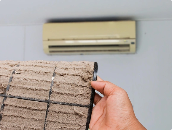 Close-up of a dirty air conditioner filter covered in dust with the unit in the background
