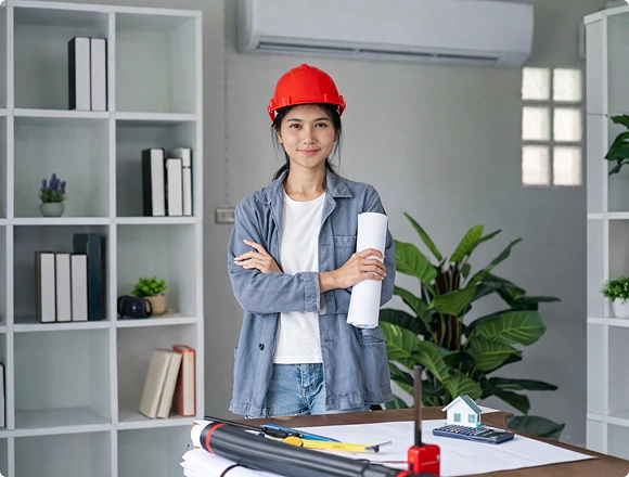 Female HVAC technician wearing a hard hat standing in front of a wall-mounted air conditioner with blueprints