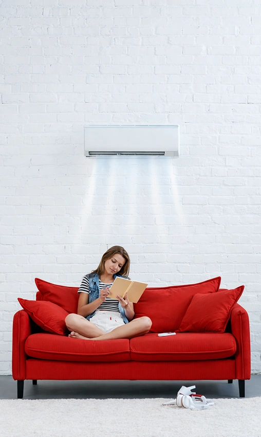 Woman sitting on a red sofa reading a book under a wall-mounted air conditioner