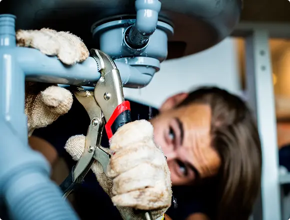 Person using pliers to tighten or repair a leaking pipe under a sink, focusing on plumbing maintenance.