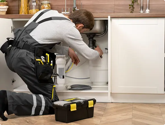Plumber working under a kitchen sink, adjusting or repairing the plumbing system with tools nearby.