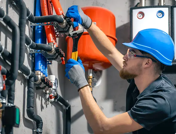 Technician wearing safety gear using a wrench to adjust or repair a network of pipes and valves in a mechanical or utility room.