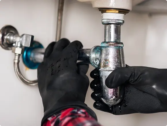 Hands in protective gloves tightening or repairing a metal drain pipe connection under a sink.
