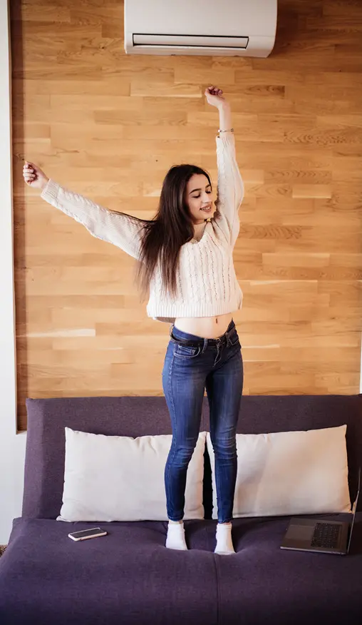 Woman standing on a couch stretching happily under a wall-mounted air conditioner, suggesting comfort and a pleasant indoor climate.