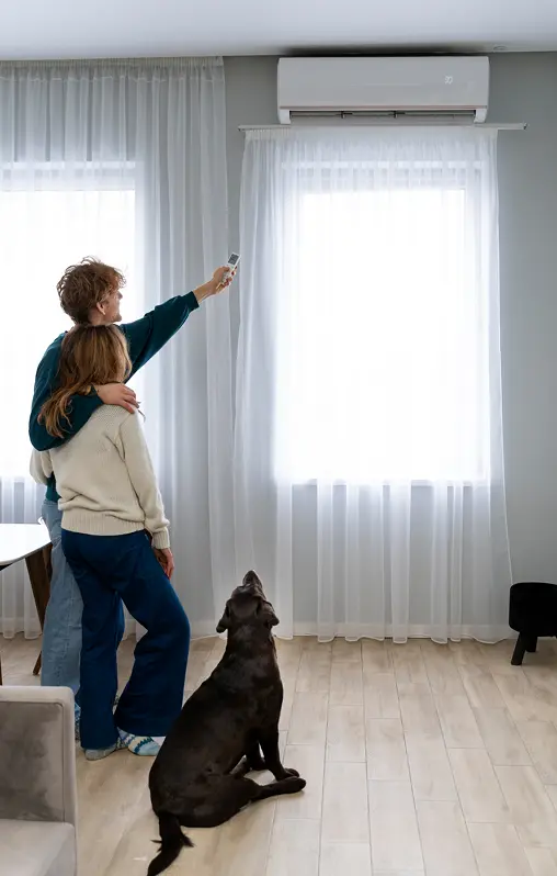 Couple standing together in a living room, adjusting an air conditioner with a remote, while a dog sits nearby.