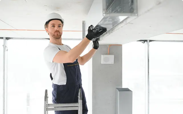 A technician in a white hard hat standing on a ladder installing a ceiling ventilation duct