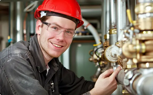 A smiling engineer in a red hard hat and safety glasses working on industrial pipeline valves and gauges