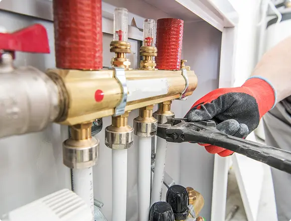 A gloved hand using pliers to adjust fittings on a brass heating manifold with flow meters and valves
