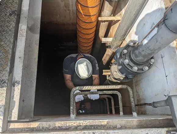 A worker wearing a hard hat descending into an underground utility shaft with large pipes and a spiral cable