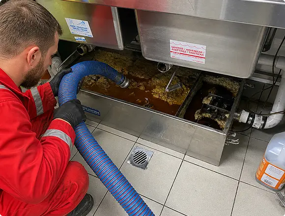 A technician in a red uniform cleaning a commercial grease trap with a suction hose in a kitchen