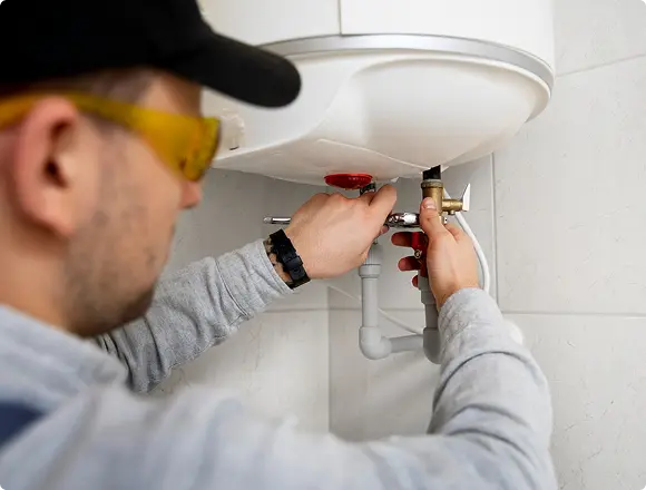 A plumber wearing yellow safety glasses adjusting the valve connections underneath a wall-mounted water heater